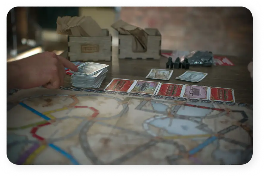 Ręka wskazuje na stos kart obok rozłożonego planszy gry „Ticket to Ride”